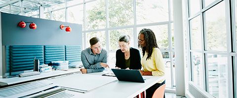 Three coworkers look at a laptop inside a conference room.
