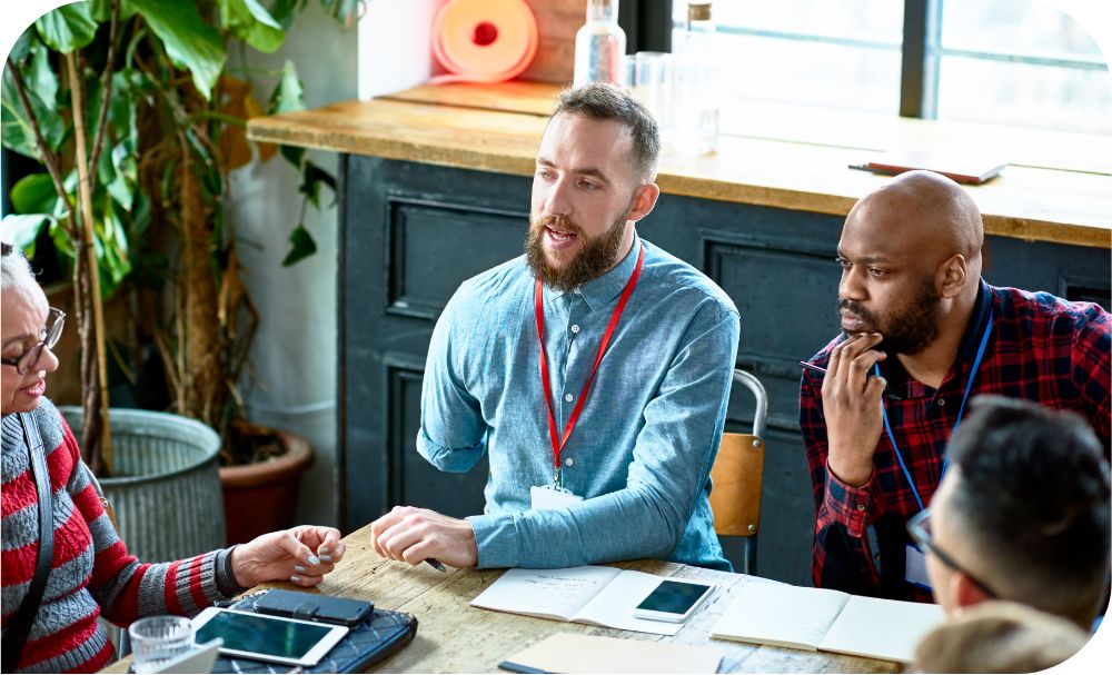 A group of people in casual business attire with badges on lanyard necklaces sit at a table and talk 