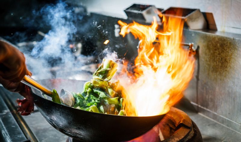 Close-up do braço de um chef segurando uma wok de ferro com vegetais e uma chama forte