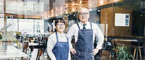 Woman and man standing in front of glass doors of their restaurant behind them.