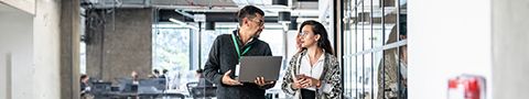 A person holding a laptop standing in an office setting talking to another person