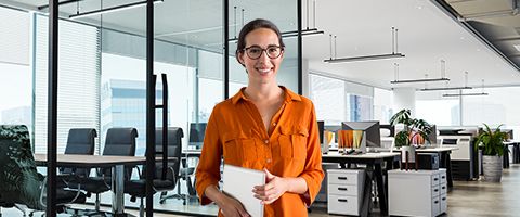 Person in an orange shirt standing and holding a smart tablet in an office setting.