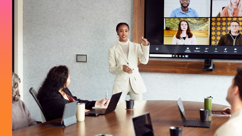 Woman in a business suit standing and presenting to a group of people sitting in a conference room and another group on screen via video conference.