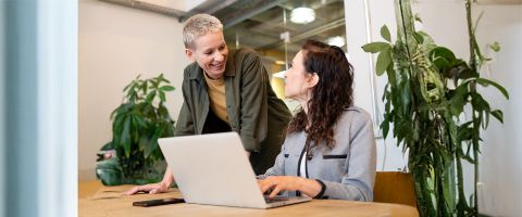 Two women in a modern office setting collaborate over a laptop.