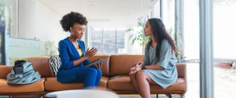 A woman in a suit speaks to another woman seated on a couch.