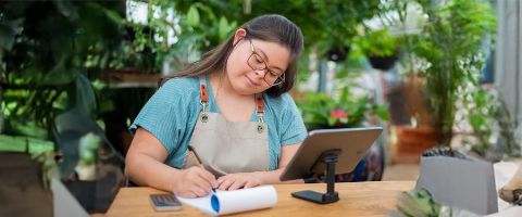 A woman wearing glasses and an apron working at a table writing in a notepad and looking at a tablet.