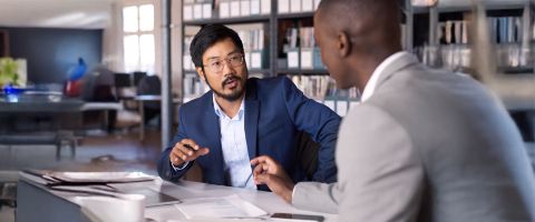 Two business professionals in suits are engaged in a discussion at a table in an office.