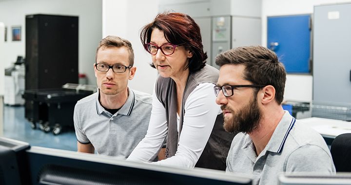 A man in casual business attire holds a tablet device while standing in a narrow hallway looking at technology behind glass windows embedded into the wall 