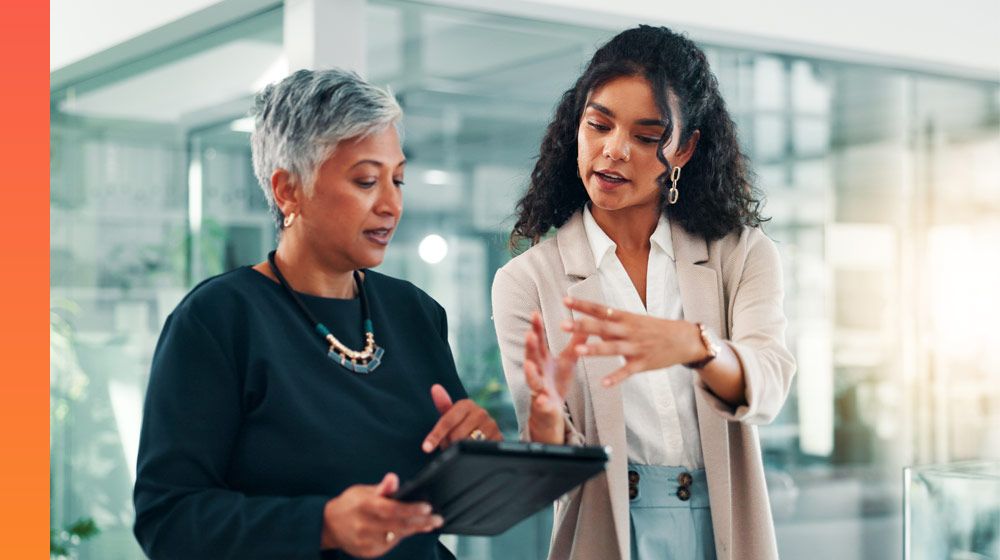 Two people in business casual attire stand in an office setting talking over a tablet.
