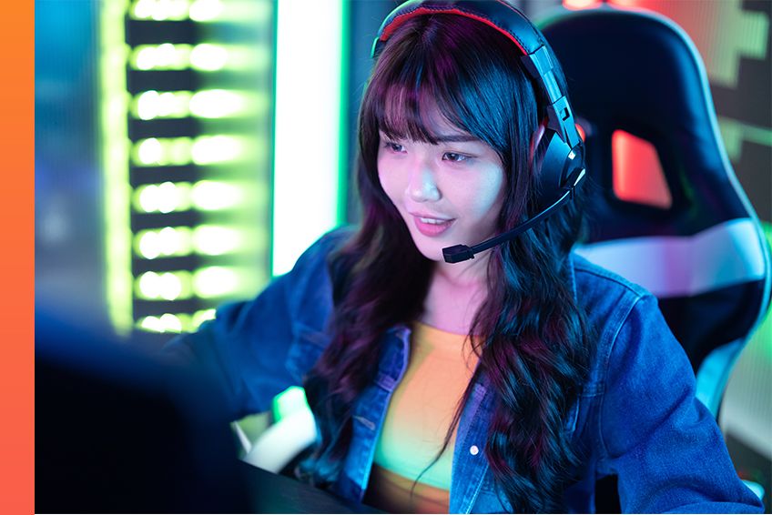 A person wearing headphones sits in a gaming chair in a colorful, tech lit room while working on the computer.