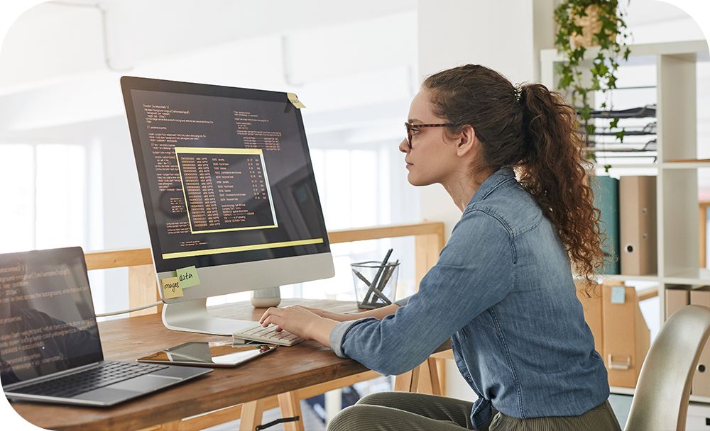 Woman sits at her desk and works from a laptop and computer monitor simultaneously 