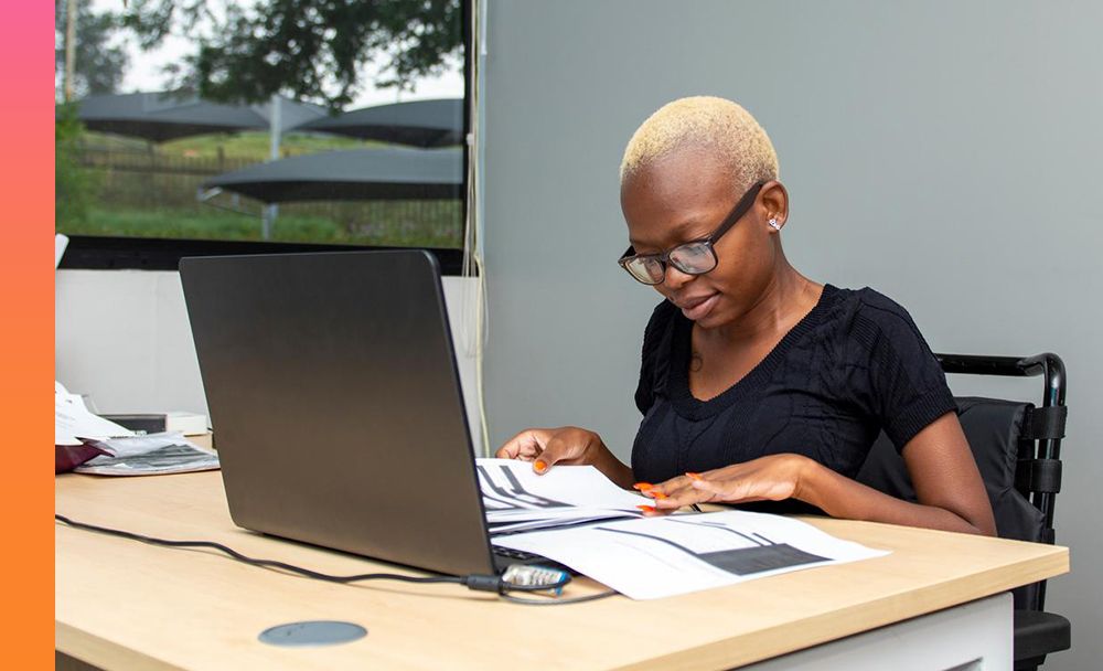 An individual in glasses and a black shirt sits at their desk reviewing a report that lays across their laptop