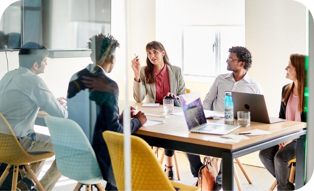 A group of business professionals sit around a table in a modern office setting with laptops and papers in front of them, having a discussion 