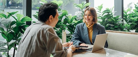 Two coworkers sit across from one another at a table with a laptop open having a conversation