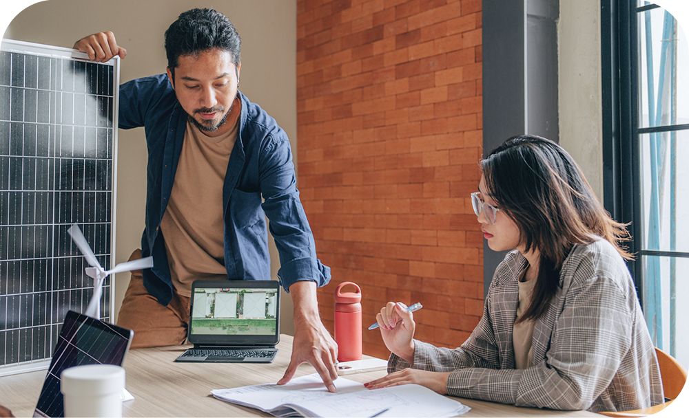 A professional stands over a desk pointing to a notebook placed in front of another professional sitting at the desk as they have a conversation