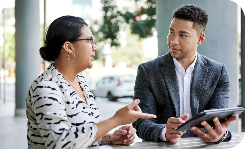 Two coworkers sit together outside and converse over a tablet