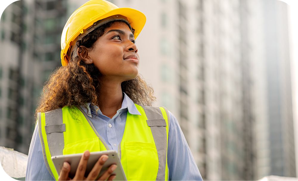 A one-armed construction worker stands outside in an orange vest and hardhat while smiling and looking at their tablet