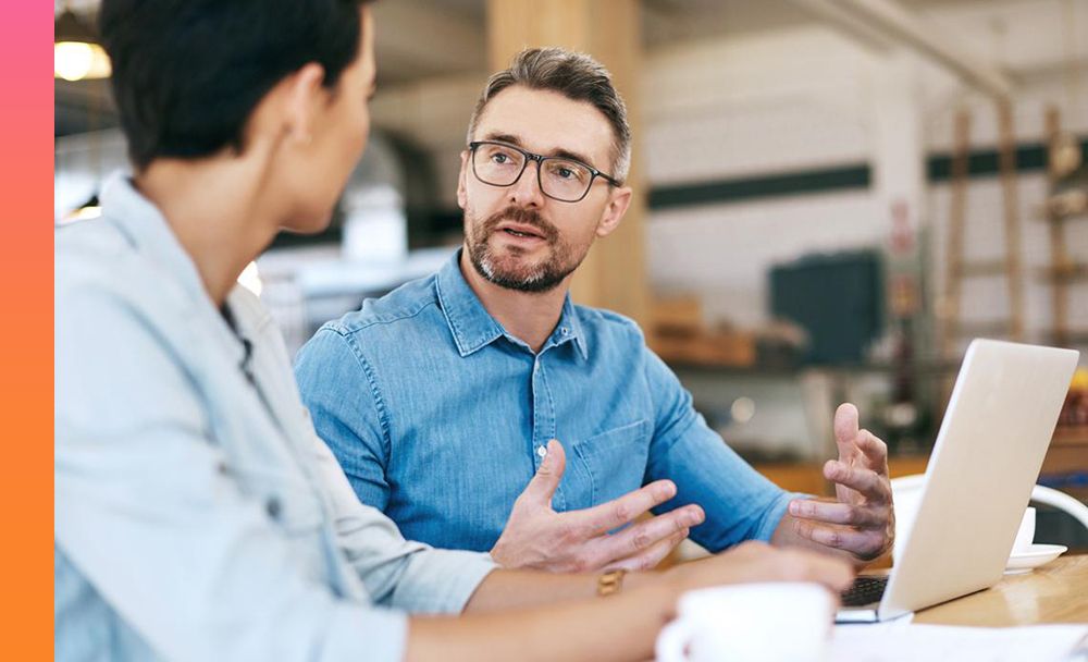 Two businesspeople sit on the same side of a table having a conversation over a laptop and coffee