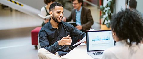 A professional sits at a table while smiling and talking about the paper in their hand to an individual across the table with a laptop in front of them