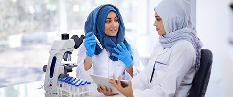 A woman holding a test tube talks with a colleague as they stand next to a table with a microscope and more test tubes 