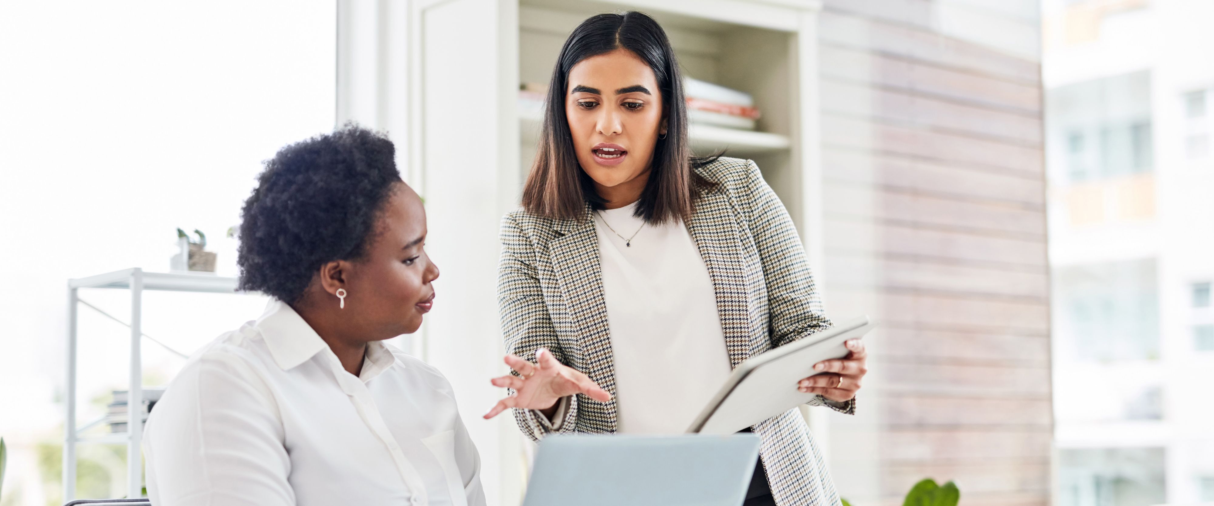 Two people in business casual attire talk over a laptop and tablet in a bright office setting.