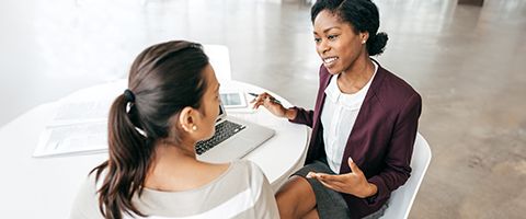 Two women sit at a table with a laptop and have a discussion.