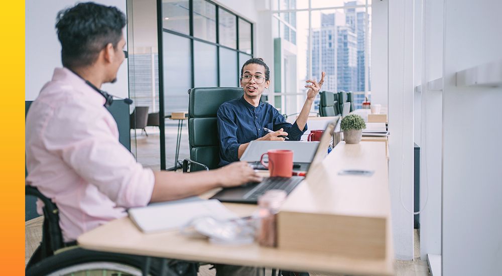 Two people siting at a row of desks while talking and working on laptop devices in front of them