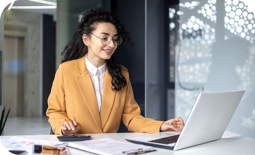 Woman in a yellow suit jacket sitting at a desk while working on a laptop device in front of her 