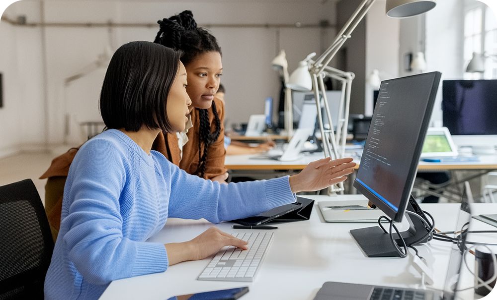Two people work together at a desktop computer in a bright modern office. One in a blue sweater sits and points at the monitor while the other leans in to look at the screen. Rows of workstations are visible in the background.