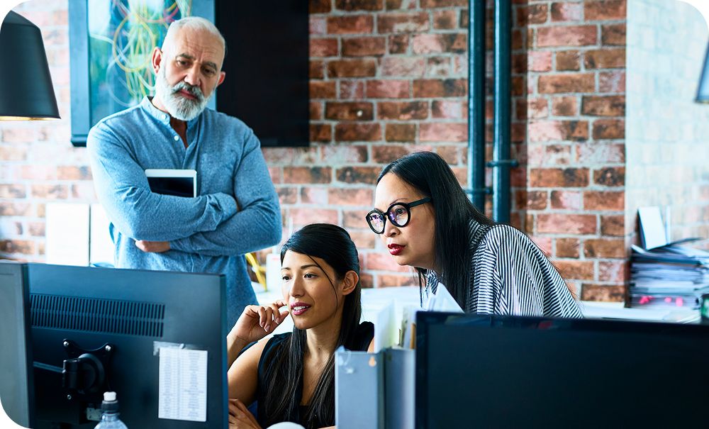 Three people collaborate around computer monitors in an office with an exposed brick wall. Two are seated and focused on the screens while a third stands behind them with arms crossed, looking toward the monitors.