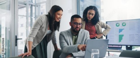 Three businesspeople collaborate around a laptop in an office while one points at a screen and a monitor in the background displays charts and graphs.
