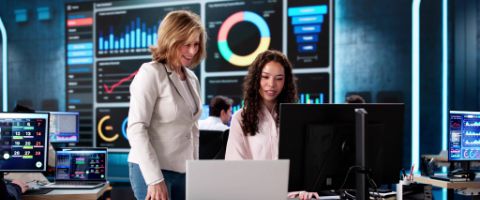 Two businesspeople work together on multiple computers in a modern office environment with large screens displaying colorful charts and graphs.