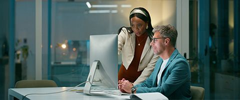 Security analyst sitting in front of monitors examining detailed threat scape data discusses action items with colleague. 