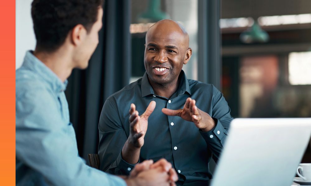 Man talking to another person sitting across from him while a laptop sits in the middle