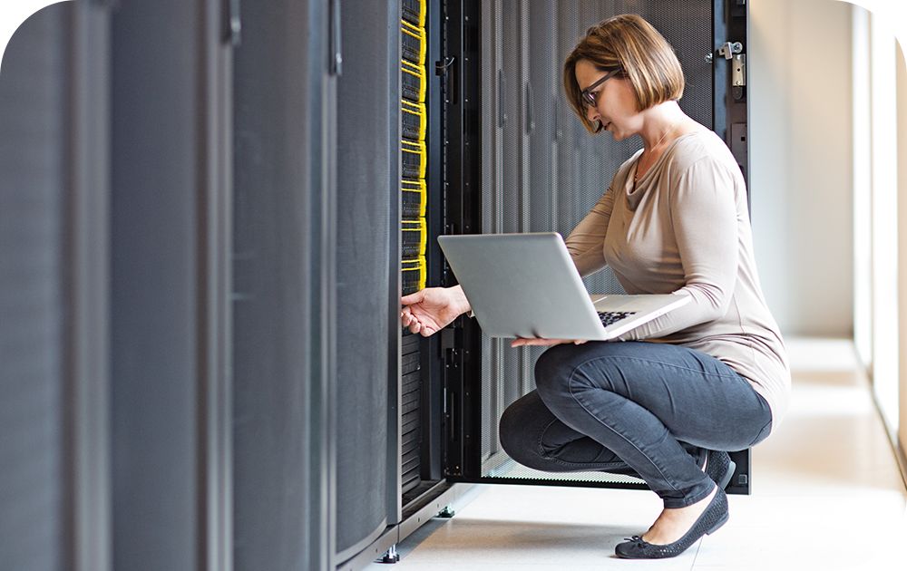 Woman kneeling in front of a server stack while adjusting it with one hand and holding a laptop in the other