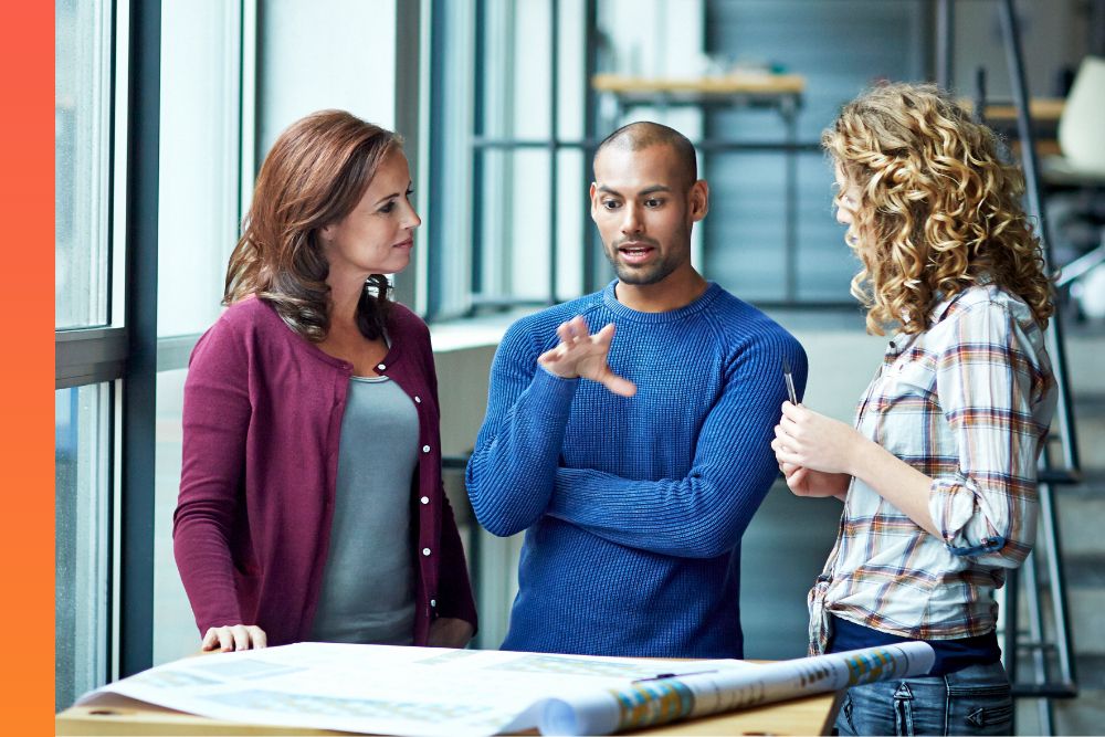 Man in a blue sweater standing while talking to two other people over prints out on a table