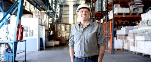 Man standing on a factory floor while wearing a hard hat with his hands in his pockets