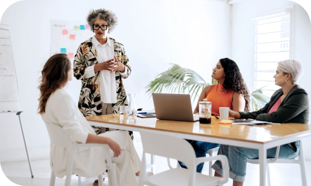 Woman standing while talking to three other people while they sit at a table with a laptop and papers