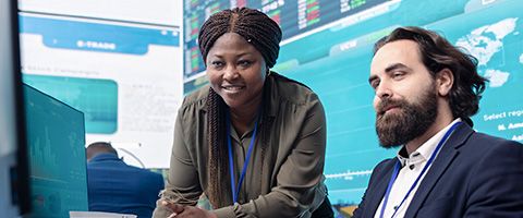 A person in business attire smiling at a computer with a colleague standing next to them. 