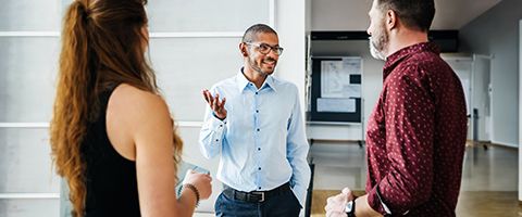 Person in business attire standing in an office setting and talking to two other people .