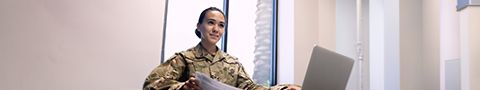 Person in military attire in a beige room working on a laptop.