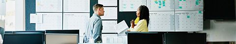 Two people standing in a classroom setting talking in front of a large board.