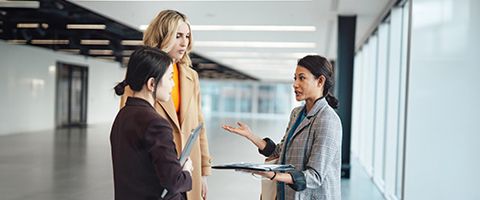 Three people in business attire standing in an office setting and having a discussion.