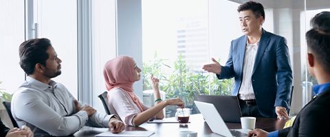 A professional stands at the head of a table and speaks to the individuals seated around them.