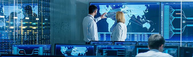 Two business people standing in front of a digital map in a room of computer monitors with digital maps