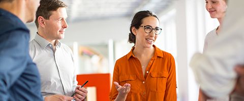  Woman in an orange shift standing while talking to other people standing around in a circle