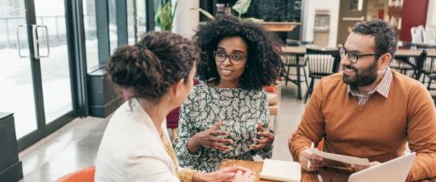 Three people have a conversation while one person takes notes at a table. 