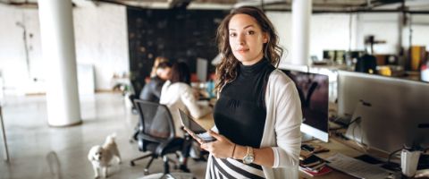 Woman in a white sweater standing while holding a tablet device in front of a row of desks