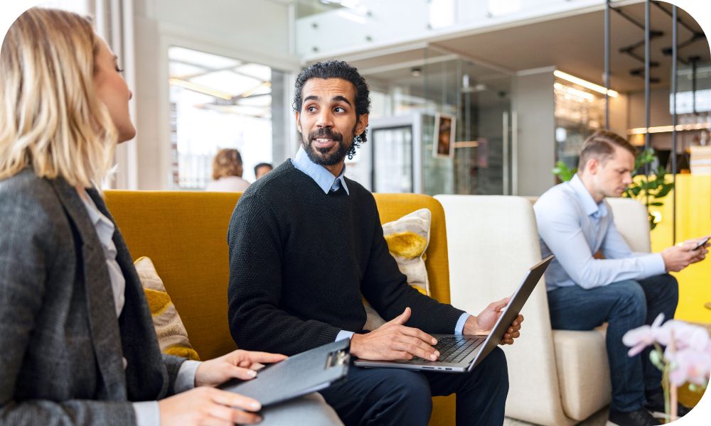 Man in a dark gray sweater sitting on a couch with a laptop while talking to a woman in a dark gray suit jacket next to him