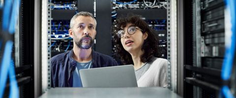 Two IT professionals in a server room looking at a laptop.